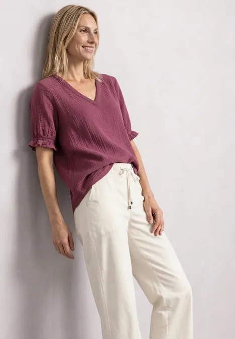 Frau lehnt entspannt an einer Wand, trägt bordeauxfarbenen Pullover und helle Hose.