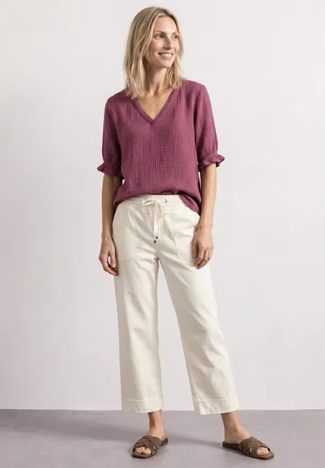 Blouse met korte mouwen, V-hals en ruches dark clouded blush Een glimlachende vrouw in bordeauxroze blouse, witte wijde broek en sandalen voor een neutrale achtergrond.