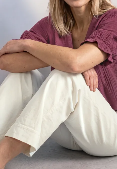 Blouse met korte mouwen, V-hals en ruches dark clouded blush Zittend persoon met gekruiste benen, draagt lichte broek en roze top, blond haar zichtbaar.