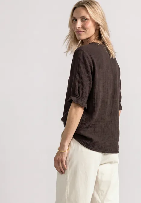 Chemisier à manches courtes, col en V et volants black coffee Femme blonde en haut noir et pantalon blanc, souriante, regard par-dessus l’épaule.