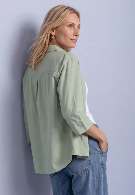 3/4 Arm Hemdbluse mit Cordmix eucalyptus green Eine Frau in hellgrüner Bluse und Jeans steht seitlich vor einer Wand und blickt nach rechts.