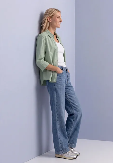 3/4 Arm Hemdbluse mit Cordmix eucalyptus green Frau lehnt lässig an Wand, trägt grünes Sakko, weißes Shirt, weite Jeans und silberne Sneaker.