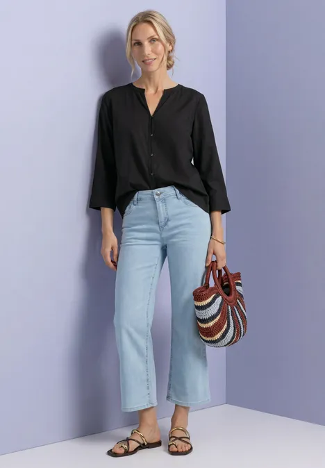 Frau mit schwarzer Bluse, hellblauen Jeans, gestreifter Tasche und Sandalen vor lila Hintergrund.