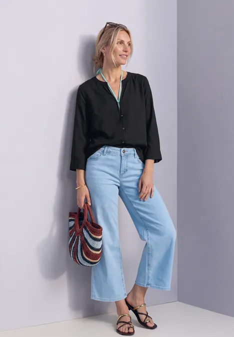 Frau lehnt an Wand, trägt schwarze Bluse, weite helle Jeans, schwarze Sandalen und bunte Streifentasche.