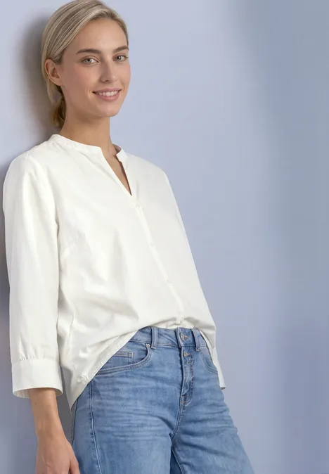 Eine junge Frau mit blonden Haaren trägt eine weiße Bluse und blaue Jeans vor hellem Hintergrund.