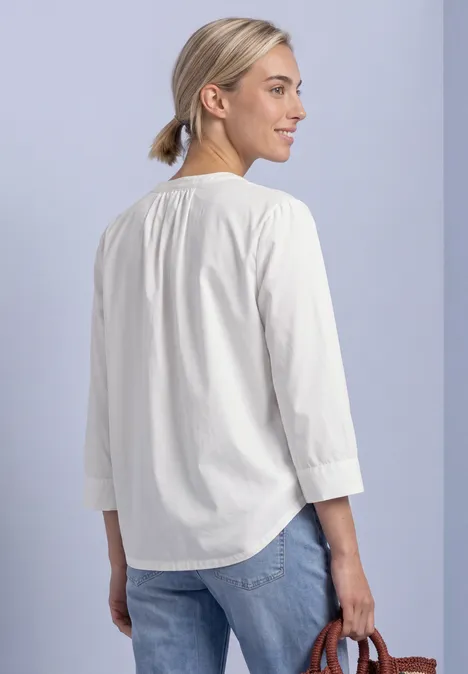 Frau von hinten in weißer 3/4-Arm-Bluse mit blondem Dutt, Jeans und kleiner brauner Tasche.