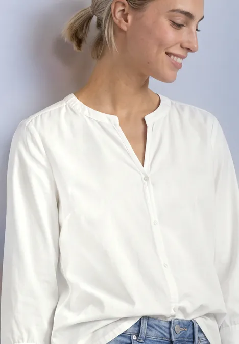 Eine lächelnde Frau trägt eine weiße Bluse mit Knopfleiste und Jeans.