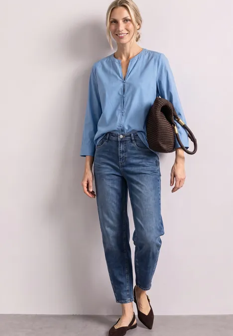 Junge Frau vor heller Wand, trägt blaue Bluse, Jeans und schwarze Ballerinas, hält eine Tasche.