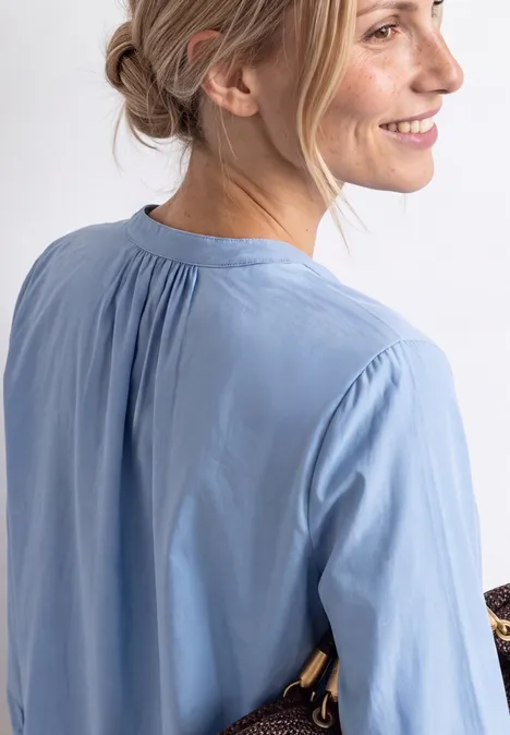Seitenansicht einer lächelnden Frau mit zusammengebundenem Haar, die eine hellblaue Bluse trägt.