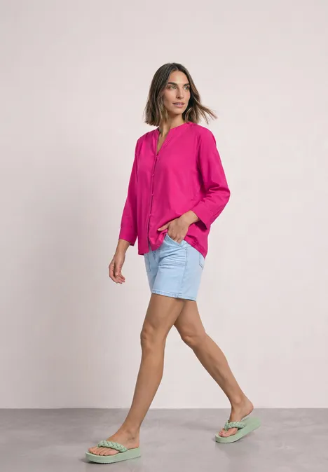 Une femme porte un chemisier fuchsia, des shorts en jean clair et des sandales vert menthe, marchant de profil.