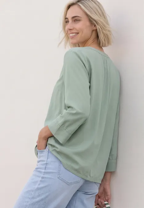 Lächelnde Frau mit kurzen blonden Haaren in grünem Langarmshirt und blauen Jeans vor heller Wand.