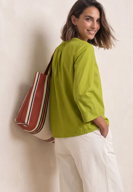 Femme souriante en chemise verte, pantalon blanc, sac rayé porté à l’épaule
