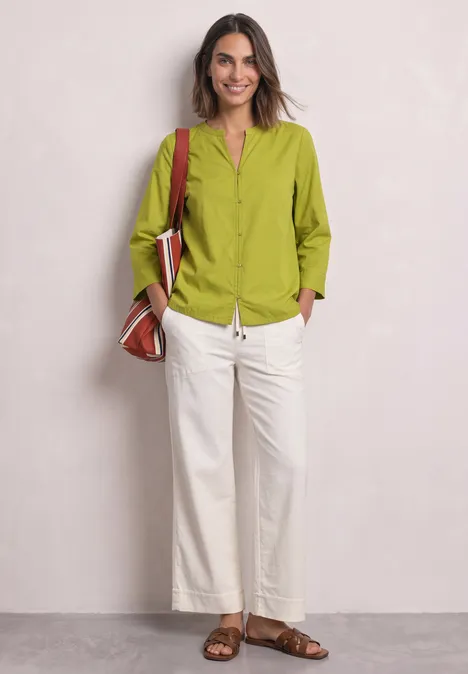 Une femme souriante pose devant un mur clair, vêtue d'un chemisier vert, d'un pantalon blanc et de sandales marron.