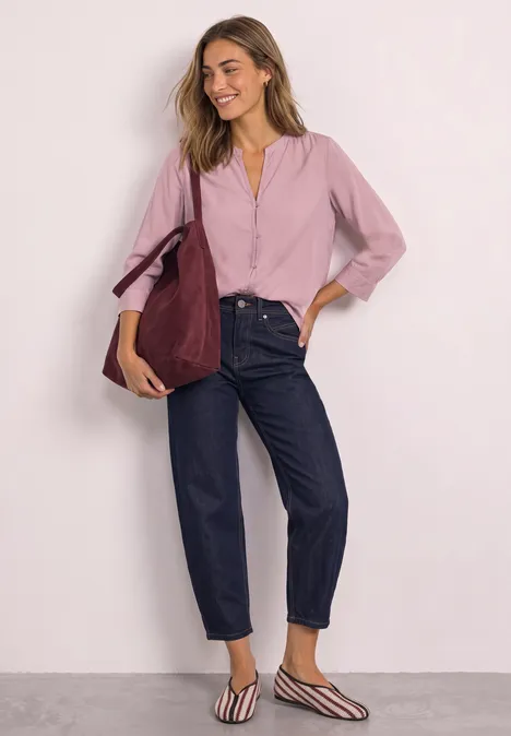 Frau lehnt lächelnd an der Wand, trägt rosa Bluse, dunkle Jeans, gestreifte Ballerinas und bordeauxrote Tasche.
