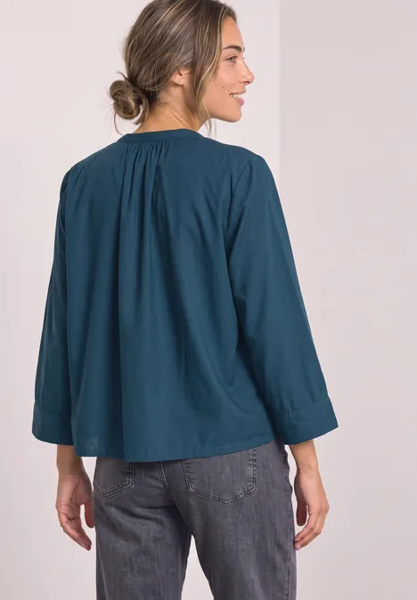 Frau in taubenblauer Bluse mit weiten Ärmeln und Jeans, von hinten fotografiert.