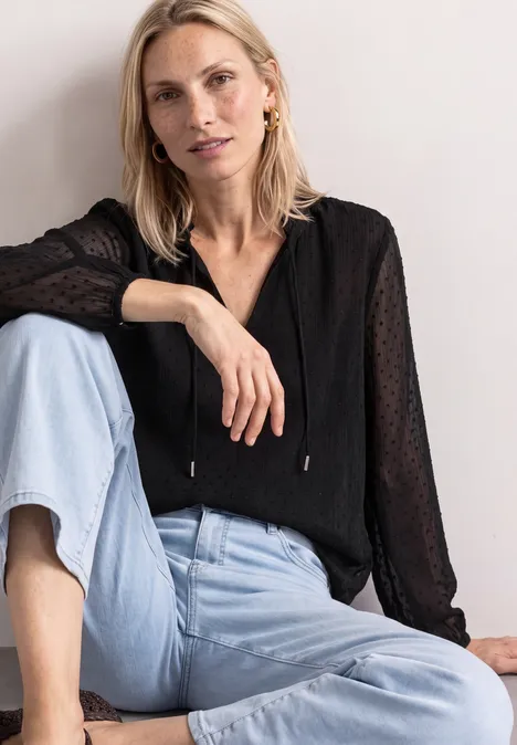 Blondynka siedzi zrelaksowana, ma na sobie czarną bluzkę i jasnoniebieskie dżinsy.