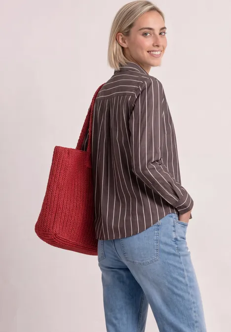 Femme en chemise rayée, jean et sac rouge, tournant la tête avec un sourire discret.