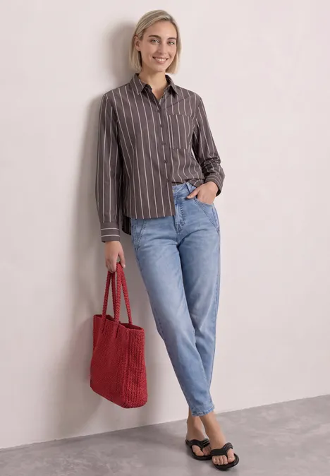 Femme appuyée contre un mur, porte une chemise rayée, jean bleu clair, sandales noires et sac rouge.