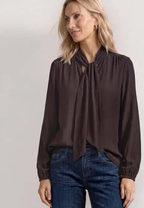 Frau trägt dunkelbraune Bluse mit Knoten am Kragen und Jeans.