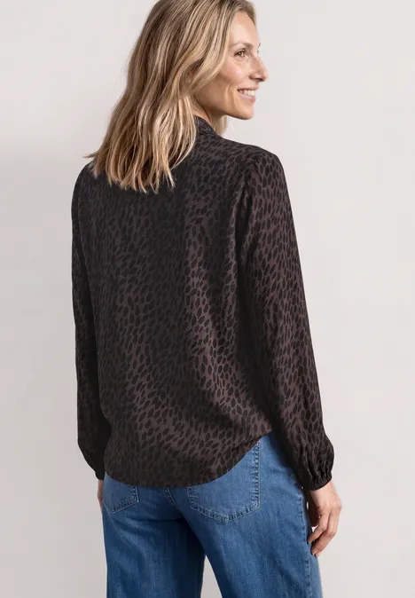 Frau von hinten in dunkelbrauner Leoprint-Bluse und blauen Jeans vor hellgrauem Hintergrund.