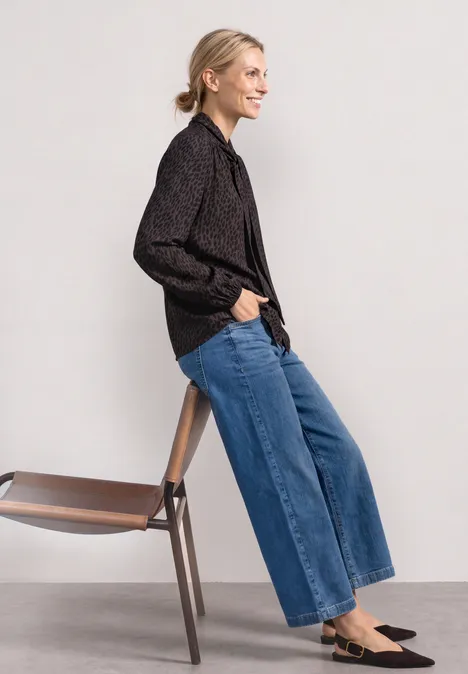 Seitlich abgebildete Person mit schwarzem Hemd, weitem Jeans, schwarzen Loafers lehnt an einem Holzstuhl.
