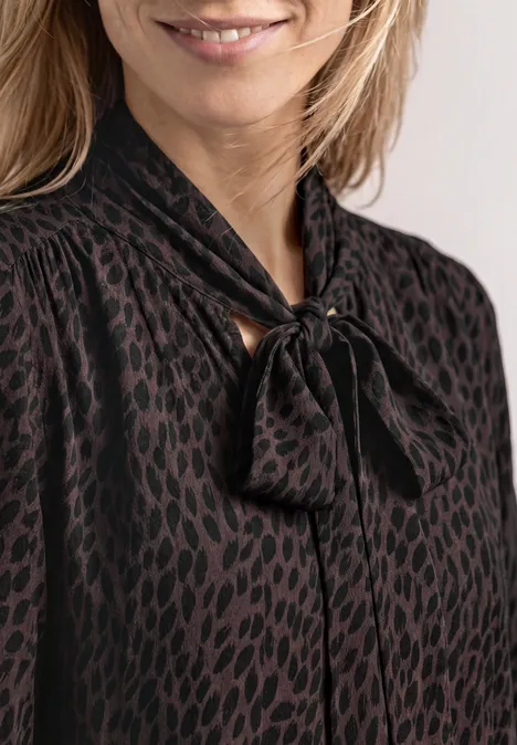 Frau in schwarzer Bluse mit dezentem Leopardenmuster und Schleifenkragen.