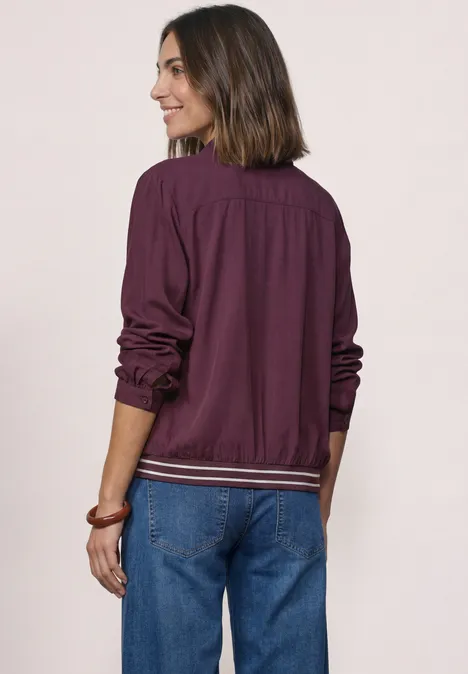 Frau mit schulterlangen Haaren, lila Jacke und blauen Jeans, lächelt und steht vor hellem Hintergrund.