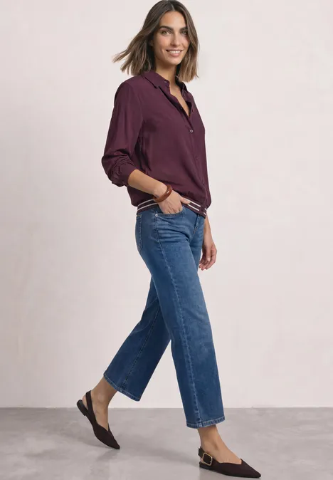 Frau in lässiger Jeans, burgunderfarbener Bluse und flachen Schuhen, die lächelnd seitlich posiert.