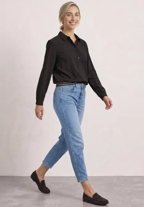 Eine lächelnde Frau in schwarzer Bluse, hoch geschnittenen blauen Jeans und braunen Loafers vor hellem Hintergrund.