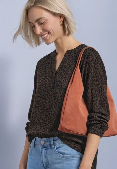 Eine Frau in gemustertem Pullover, Jeans und braun-orangener Tasche lächelt und blickt nach unten.