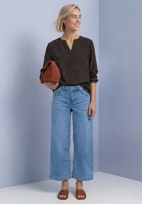 Frau vor neutralem Hintergrund in schwarzer Bluse mit Tupfen, weitem Jeans, braunen Sandalen und roter Clutch.