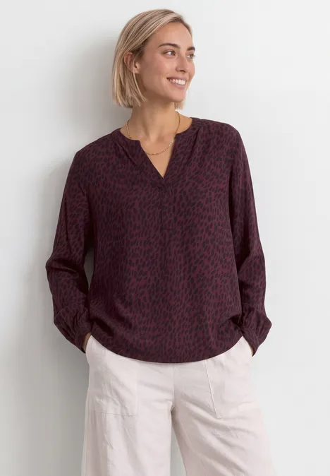 Eine lächelnde Frau mit kurzen blonden Haaren trägt eine weinrote gemusterte Bluse und helle Hose.