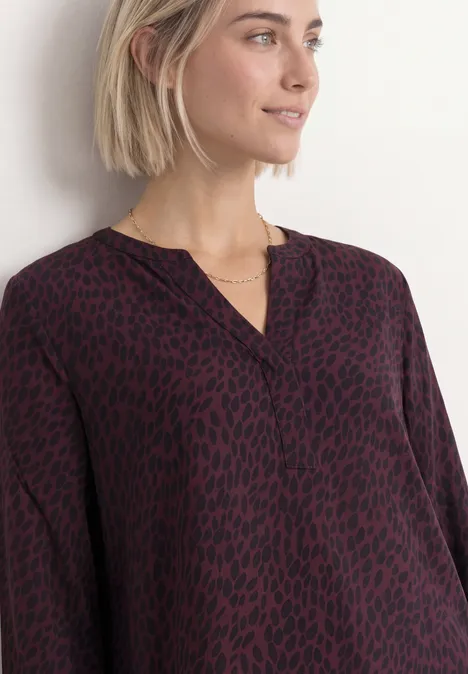 Frau mit kurz blondem Haar trägt locker geschnittene, dunkelrotschwarze Bluse mit V-Ausschnitt und Animalprint.