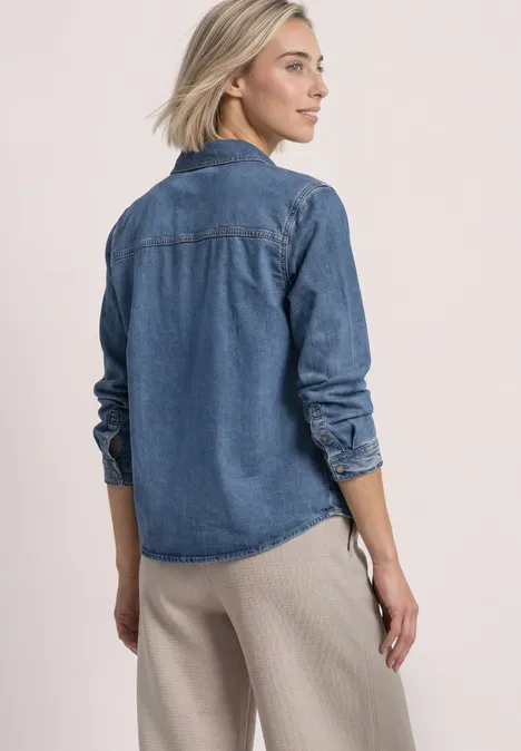 Frau im Profil mit blauer Jeansbluse und beiger Hose vor neutralem Hintergrund.