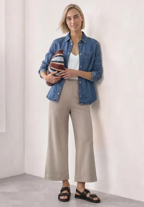 Frau in lässigem Outfit aus Jeanshemd, weißem Top und beigen Hosen hält einen gestreiften Ball und lächelt.