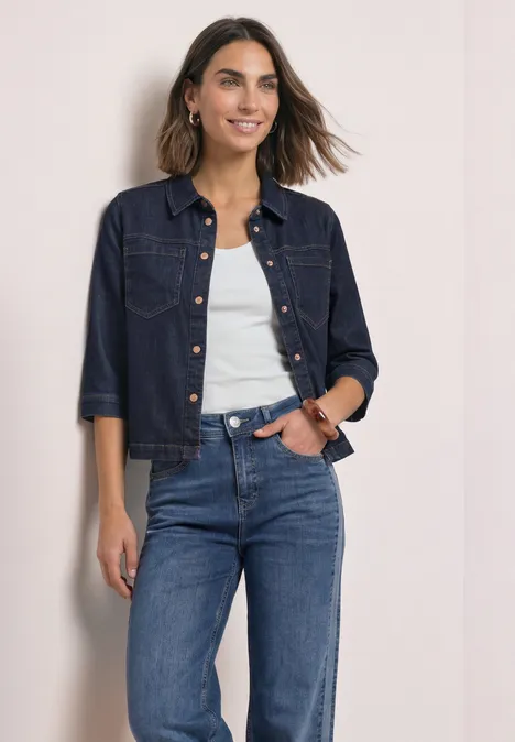 Eine lächelnde Frau in Jeansjacke, weißem Top und blauen Jeans vor hellem Hintergrund.