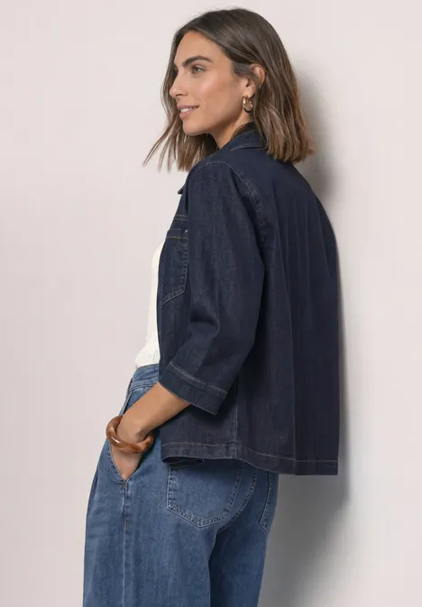Junge Frau in Denim-Jacke und Jeans, seitlich stehend und lächelnd, vor neutralem Hintergrund.