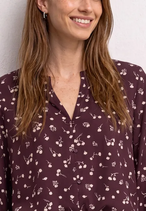 Lächelnde Frau mit langen braunen Haaren trägt eine dunkelrote Bluse mit zartem weißen Blumenmuster.