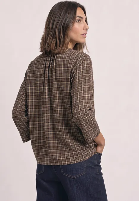 Eine Frau steht im Profil, trägt karierte braune Bluse mit langen Ärmeln und blaue Jeans.