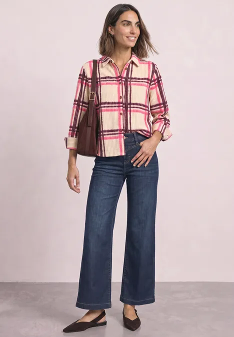 Frau in karierter Bluse, weiten Jeans und schwarzen Ballerinas trägt eine braune Schultertasche und steht lässig.