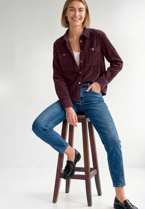Eine Frau sitzt auf einem Hocker, trägt Jeans, weiße Bluse, bordeauxfarbenes Cordjacke und schwarze Slipper.