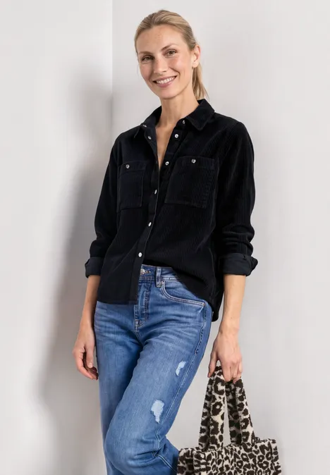 Frau in schwarzem Hemd, Jeans und Leoparden-Tasche, lächelnd vor hellem Hintergrund.