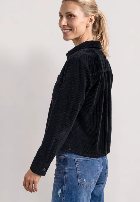 Eine Frau im schwarzen Cord-Jacket und blauer Jeans lächelt seitlich in die Kamera.