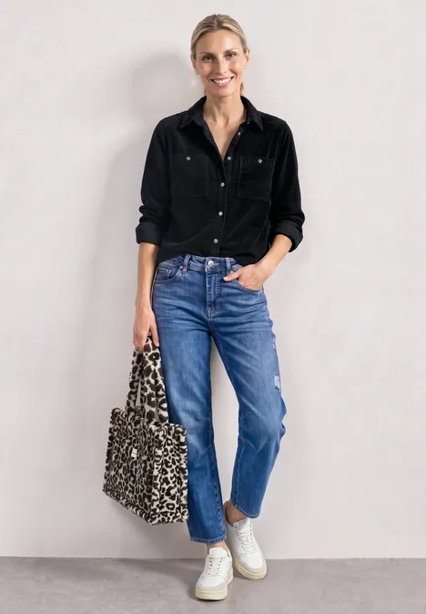 Eine Frau in schwarzem Hemd, blauen Jeans und weißen Sneakern, hält eine Leo-Tasche, lehnt an weißer Wand.