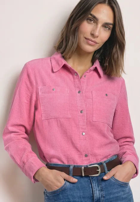 Cord-Hemdbluse mit Brusttaschen warm blush Frau in pinkem Hemd und Jeans, lächelnd mit Händen in den Hosentaschen vor hellem Hintergrund.