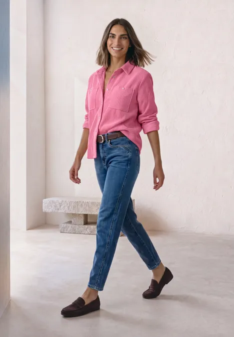 Cord-Hemdbluse mit Brusttaschen warm blush Eine Frau in rosa Bluse und blauen Jeans läuft elegant durch einen hellen, minimalistischen Raum.