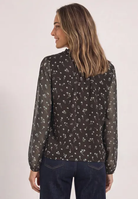 Vrouw van achteren in zwarte, transparante blouse met witte bloemenprint en blauwe spijkerbroek.