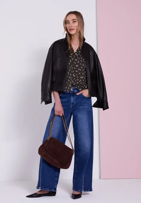 Vrouw voor een muur, zwarte leren jas op de schouders, blouse met print, wijde blauwe jeans en bruine tas.