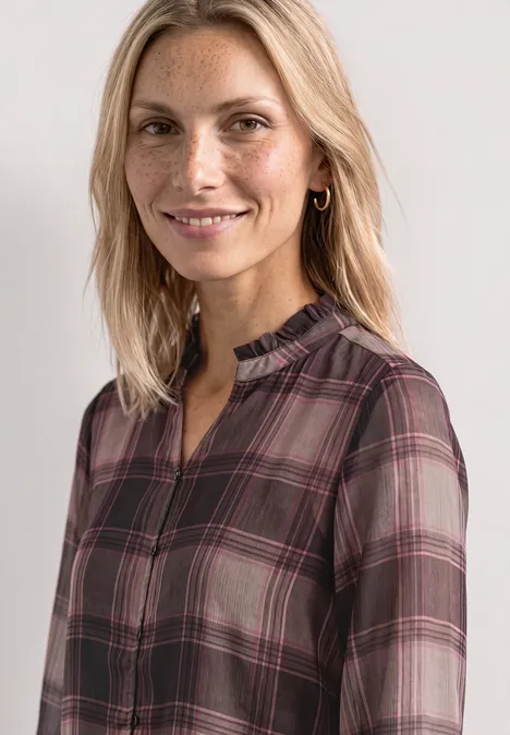 Lächelnde Frau mit blonden Haaren und kariertem Oberteil vor hellem Hintergrund.
