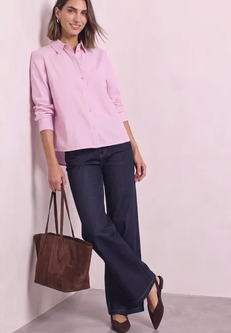 Chemisier Oxford avec imprimé dans le dos vintage blush Frau trägt rosa Hemd, weite Jeans und braune Tasche, steht lässig an einer Wand.
